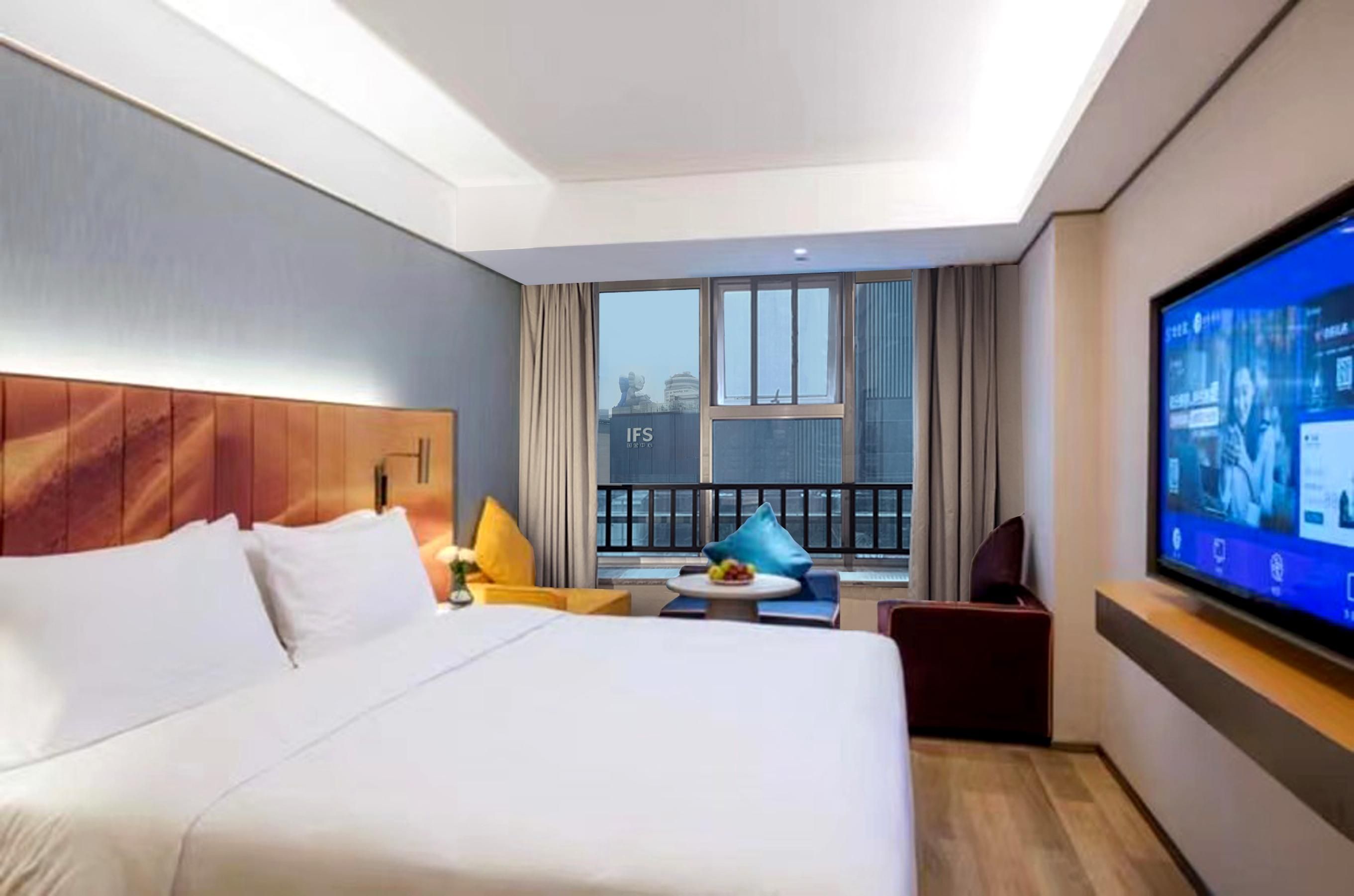 A hotel room with a large bed, a television, a balcony with curtains, and a view of the cityscape.