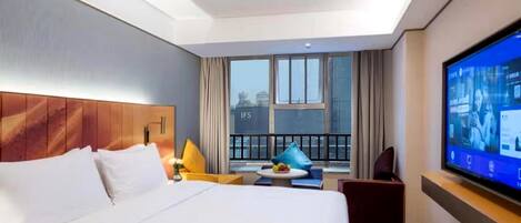 Ein Hotelzimmer mit einem großen Bett, einem Fernseher, einem Balkon mit Vorhängen und Blick auf die Stadt.