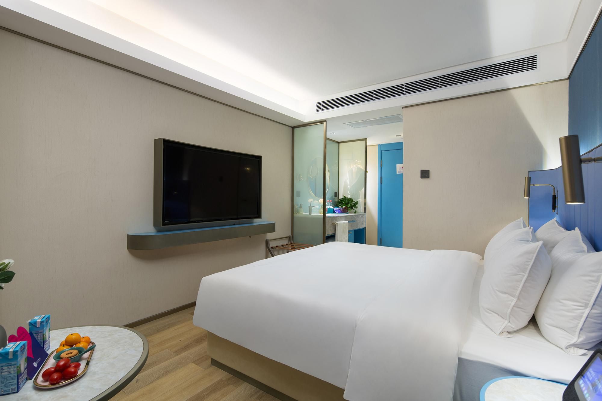 A hotel room with a bed, a flat-screen TV mounted on the wall, a round table with fruits and a bottle, and a bathroom visible through a glass door.