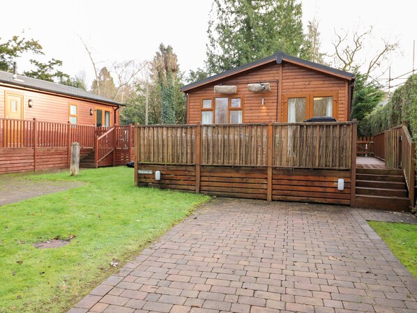 A wooden cabin with a fenced deck, a brick pathway, and a grassy yard. - Bowness Bay Lodge (Windermere)