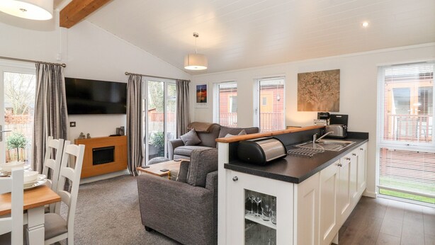A modern living room with a sofa, a dining table, and a kitchen area with a built-in oven and a coffee machine. - Bowness Bay Lodge (Windermere)