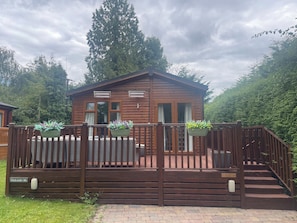 A wooden cabin with a deck, surrounded by greenery. - Bowness Bay Lodge (Windermere)