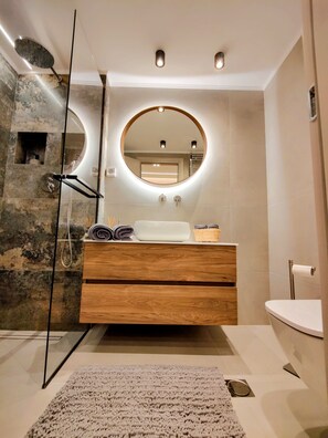 A modern bathroom with a wooden vanity, a round mirror with underglow lighting, a glass shower enclosure, and a beige tiled floor. - Elpis' cozy & luxury apartment (Egaleo)