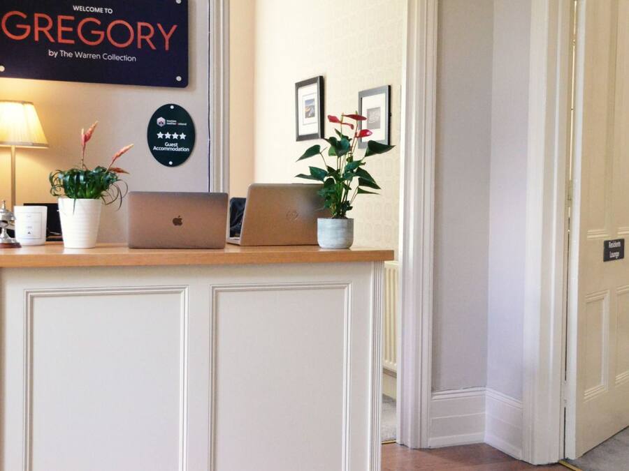 Un mostrador de recepción con dos computadoras portátiles, un letrero de alojamiento para huéspedes y un cartel de bienvenida para Gregory de The Warren Collection.