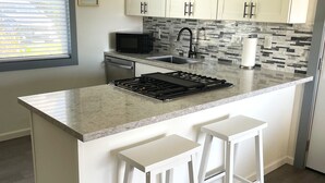 Una cocina con armarios blancos, encimera de granito, microondas negro, un lavabo y dos taburetes blancos.
