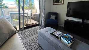 Una sala de estar con sofá, reposapiés y una mesa de centro con libros. Hay un televisor sobre un mueble bajo y un cuadro en la pared.