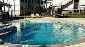 Una piscina con sillas de descanso y sombrillas, rodeada de palmeras y un edificio de fachada blanca.
