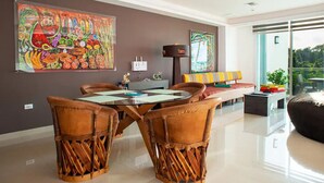 Uma área de refeições moderna com mesa de vidro e cadeiras trançadas marrons, grandes quadros abstratos nas paredes e vista para o verde lá fora.