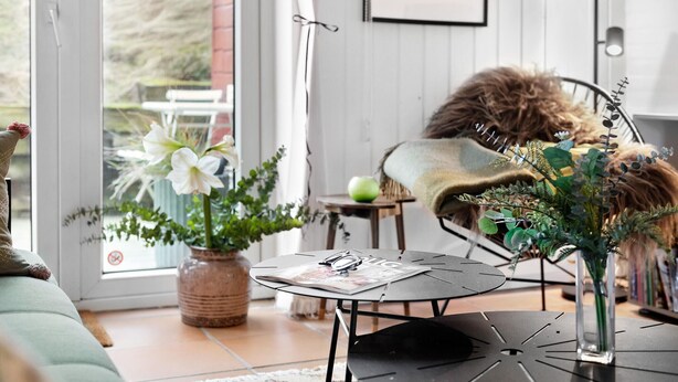 A cozy living room with a sofa, a coffee table with a magazine, a chair with a fluffy cushion, and a window with a view of greenery. - Nice home in Fanø with WiFi (Fanø)