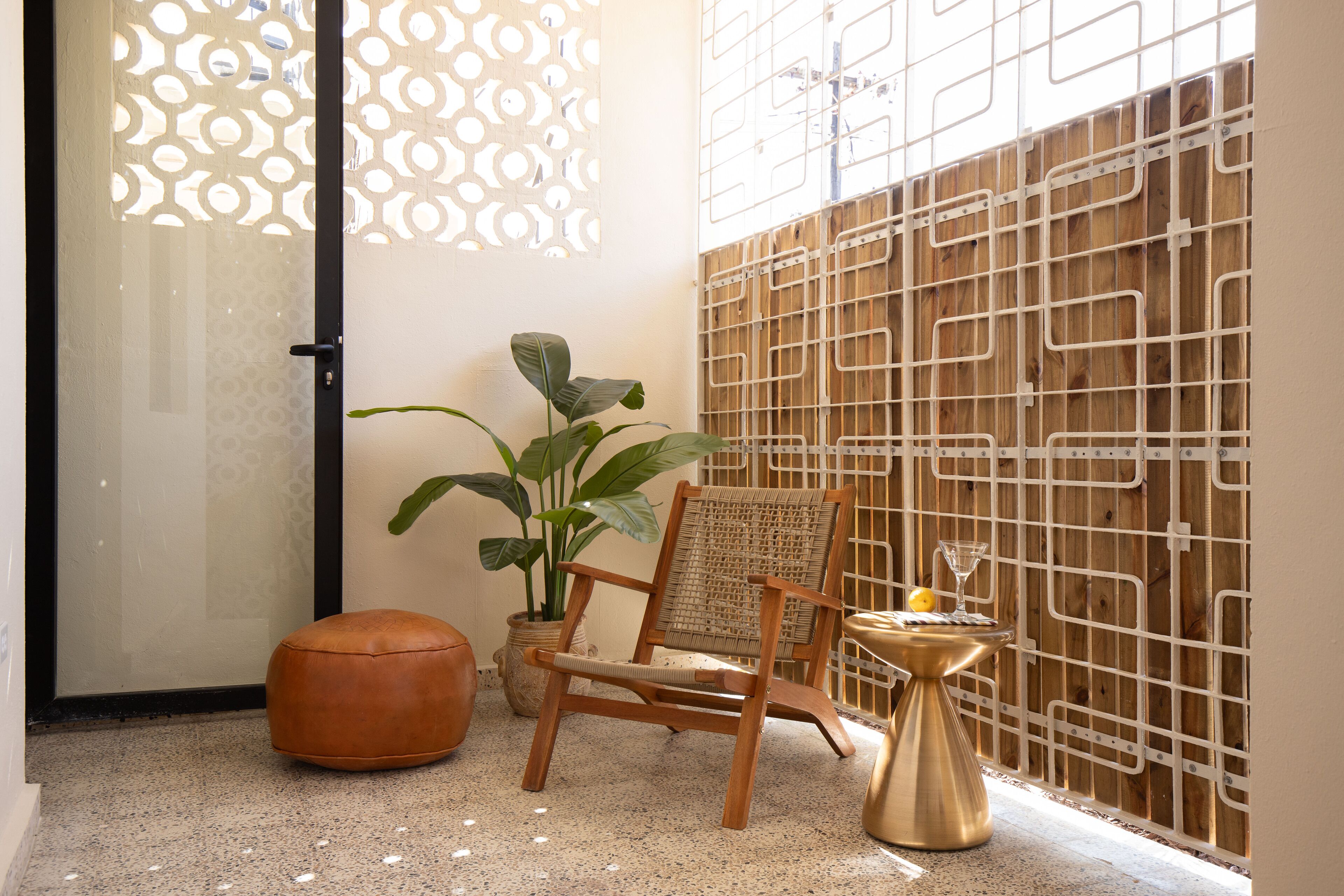 Una habitación con una silla de madera, una planta en maceta, una pequeña mesa redonda con un vaso y un panel decorativo en la pared.