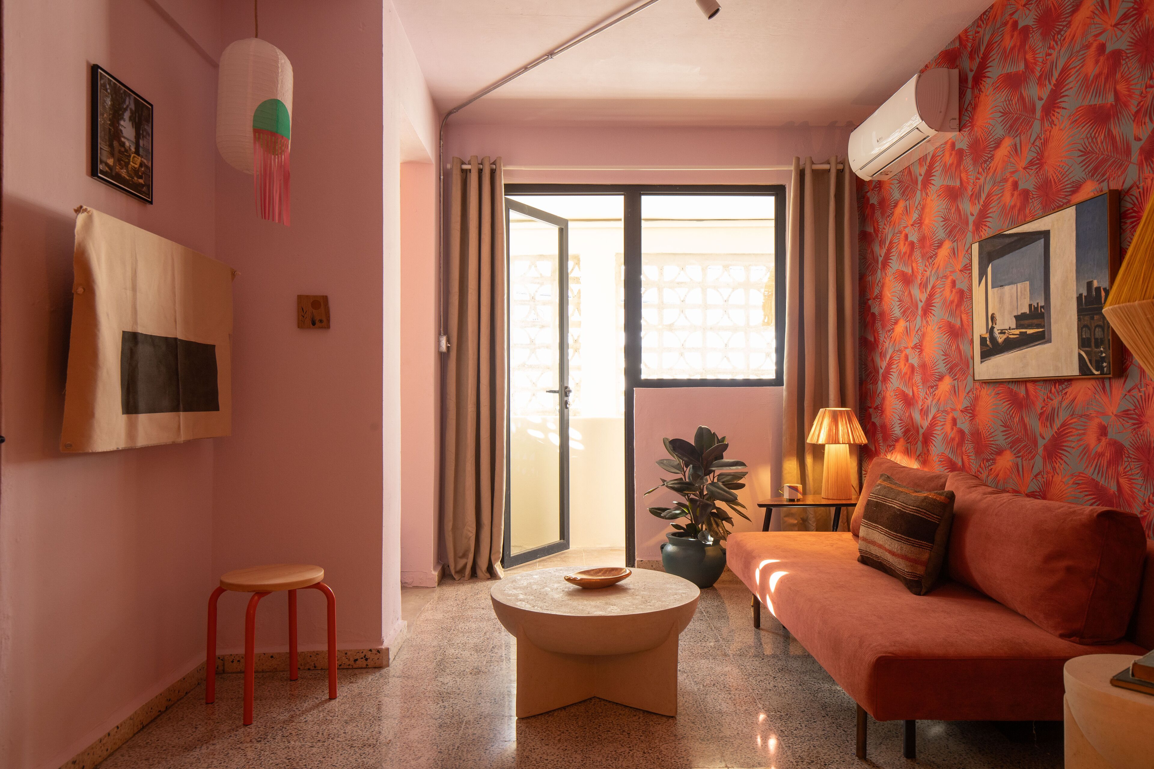 Una sala de estar con un sofá, una mesita redonda, un taburete rojo, una planta en maceta y un ventanal corredero que da a un balcón.