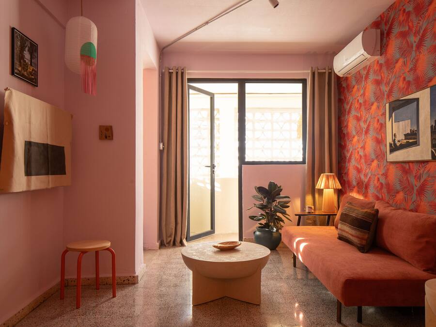 Una sala de estar con un sofá, una mesita redonda, un taburete rojo, una planta en maceta y un ventanal corredero que da a un balcón.