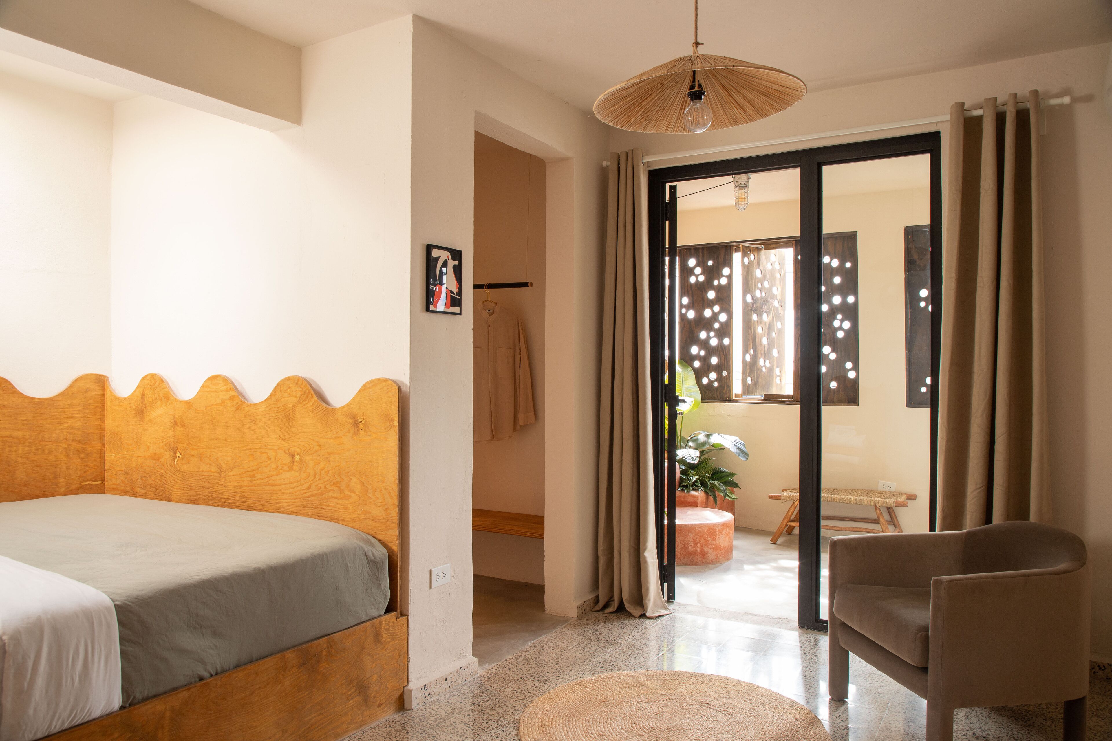 Une chambre à coucher avec une tête de lit en bois, un lit, une chaise et une porte-fenêtre donnant sur un balcon agrémenté de plantes.