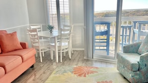 Un salon avec un canapé, une table à manger et des chaises, un tapis coloré et une vue sur l’extérieur grâce à de grandes fenêtres.