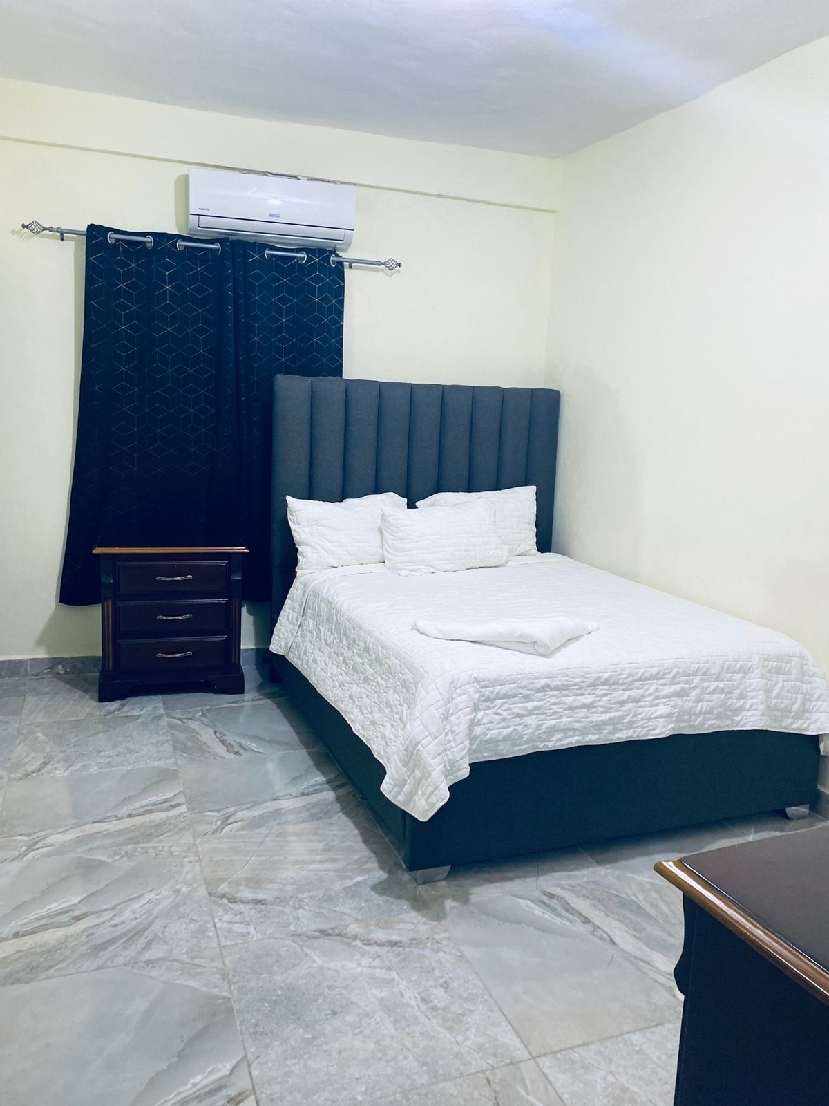 A bedroom with a bed, a nightstand, a dresser, a curtain, and an air conditioning unit.