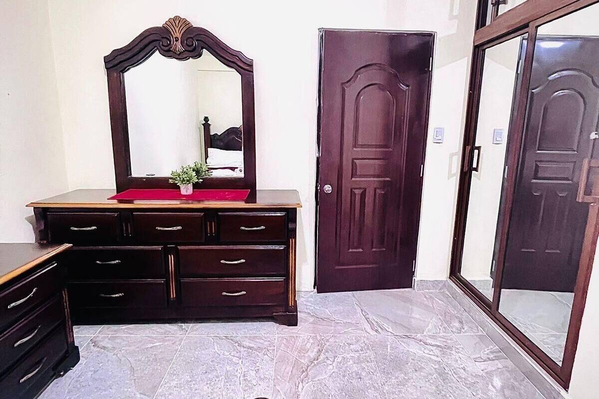 A bedroom with a wooden dresser, a mirror, a door, and a large window.