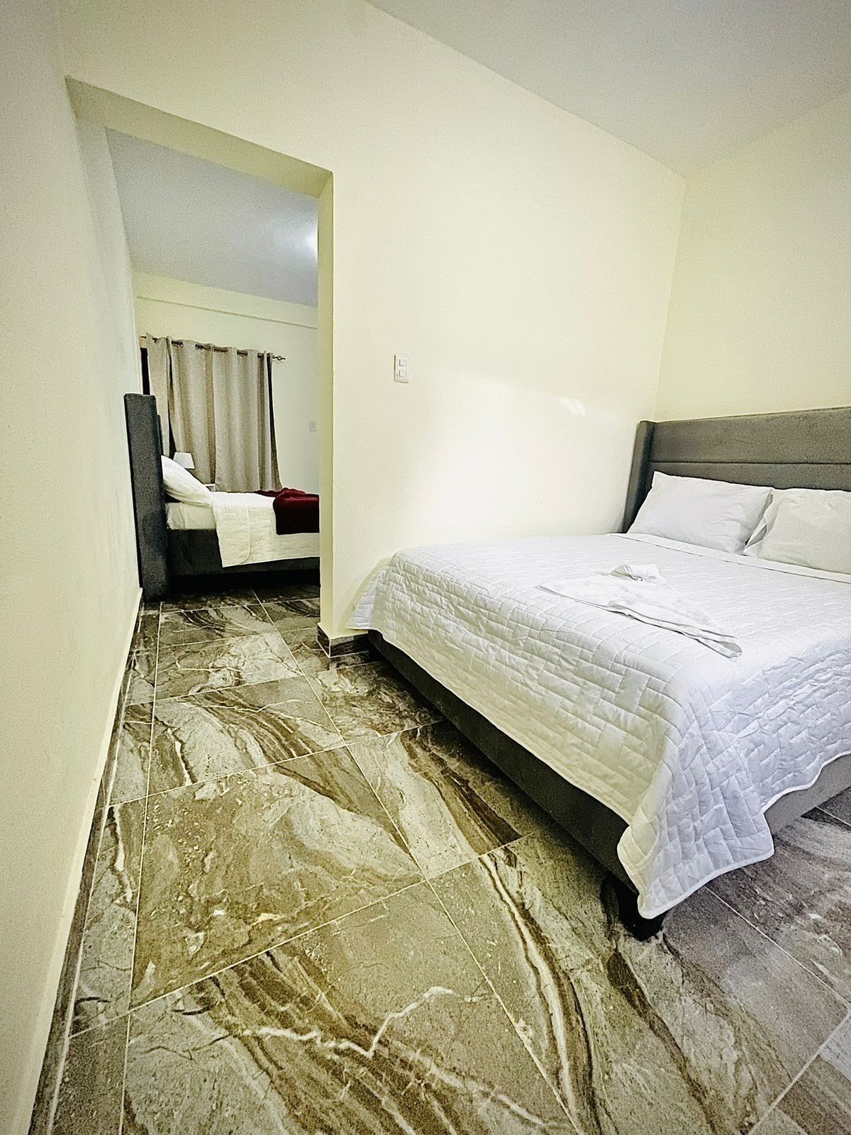 A hotel room with a marble floor, a bed with white bedding, and a view of another room through an archway.