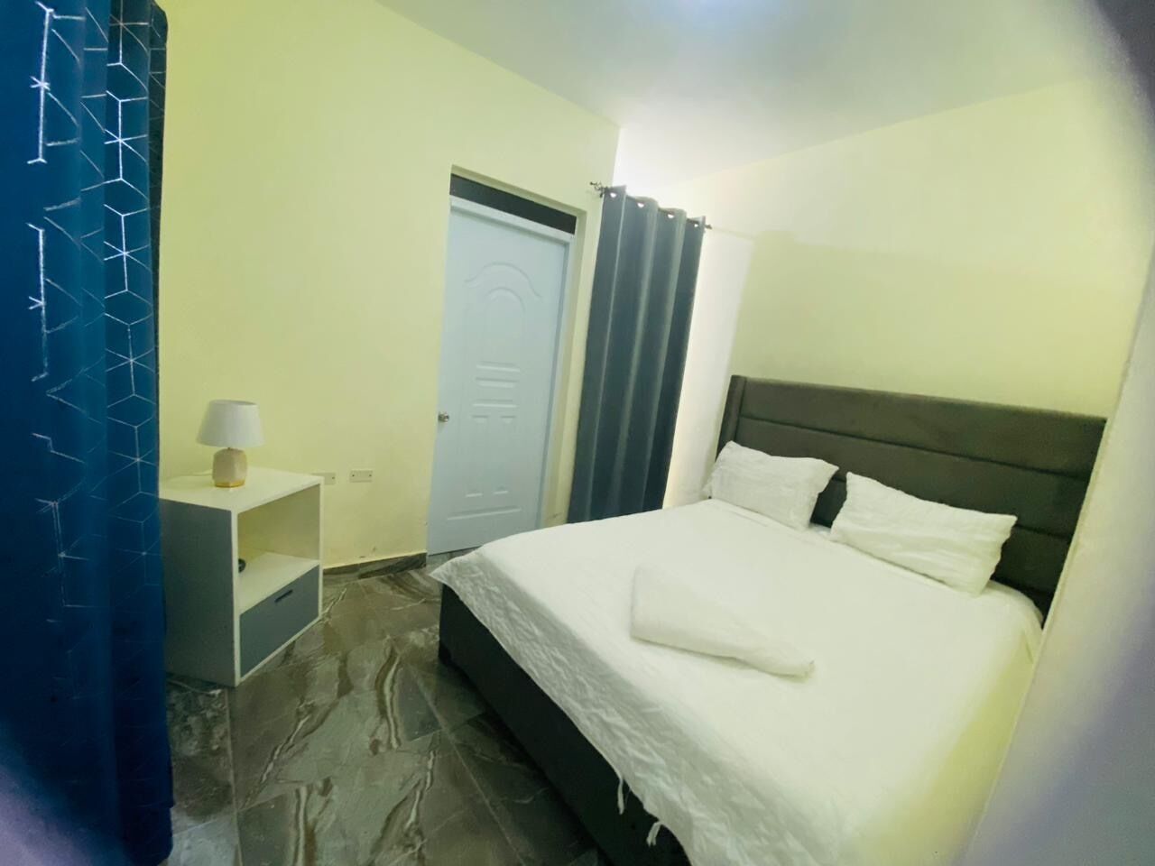 Une chambre d’hôtel avec un lit, une table de chevet, une lampe, une porte et un rideau.