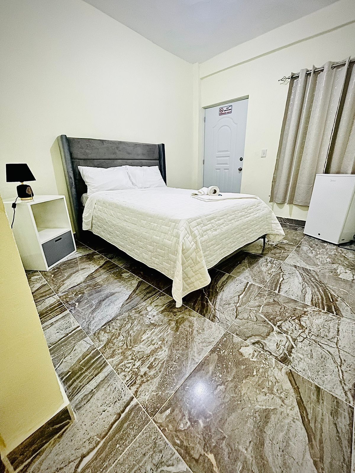 A hotel room with a bed, a nightstand, a desk, a door, and a refrigerator.