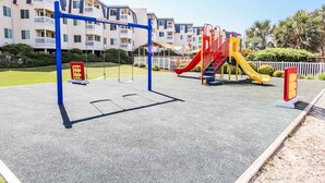 A playground with swings and slides, surrounded by a building with balconies. - Rita’s Rejuvenating Oceanfront- Pool- Hot tub- Corner unit-Sleeps 6- APATB  (Atlantic Beach)