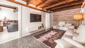 Una sala de estar con techo de madera, un televisor de pantalla plana montado en la pared, un sofá blanco, un tapete estampado y una mesita de noche con una lámpara.