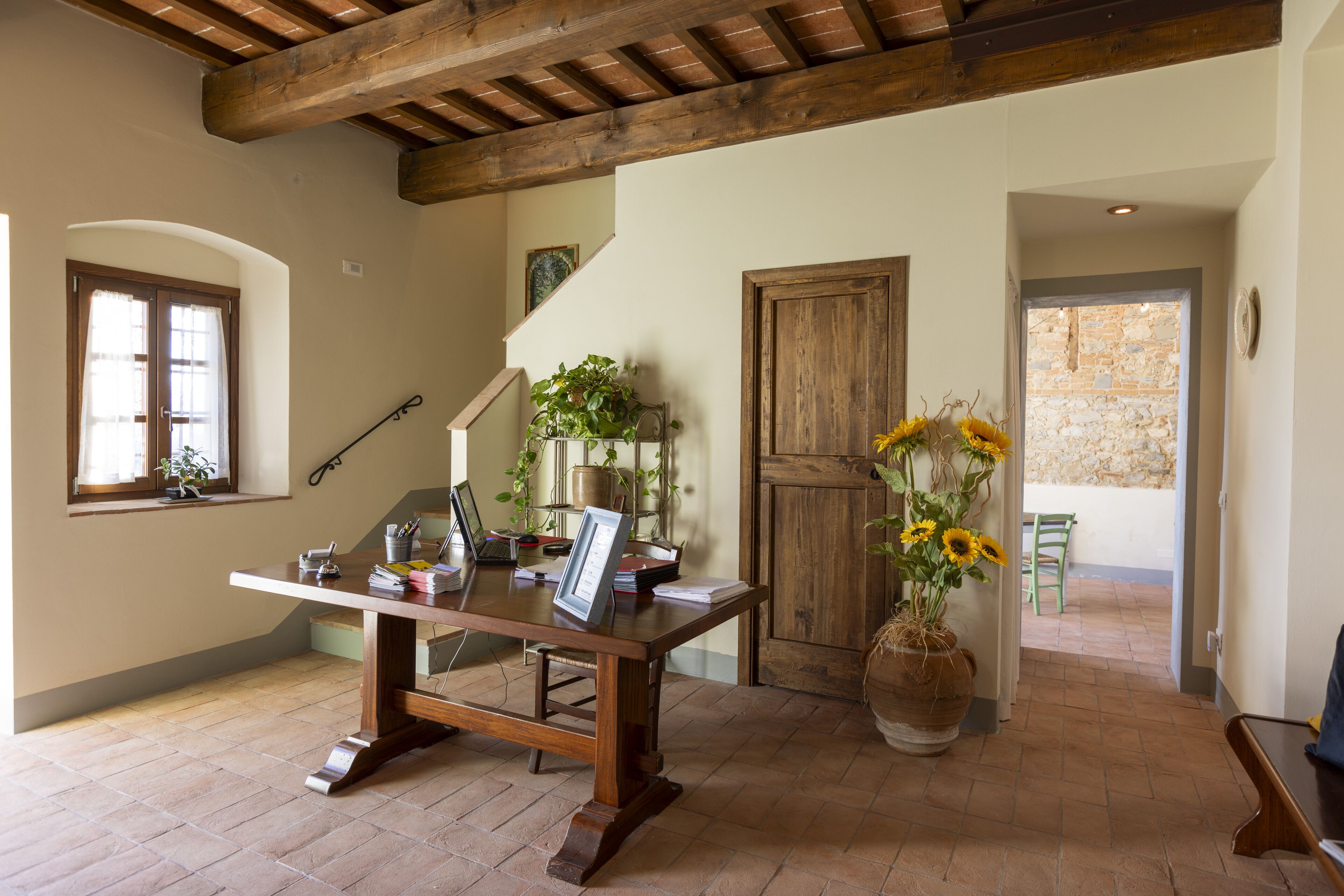 Une pièce avec une table en bois, un grand vase rempli de tournesols, un escalier et un mur de pierre visible à travers une porte ouverte.