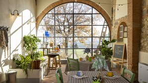 A dining area with a large arched window, checkered tablecloths, green chairs, potted plants, and a chalkboard menu.