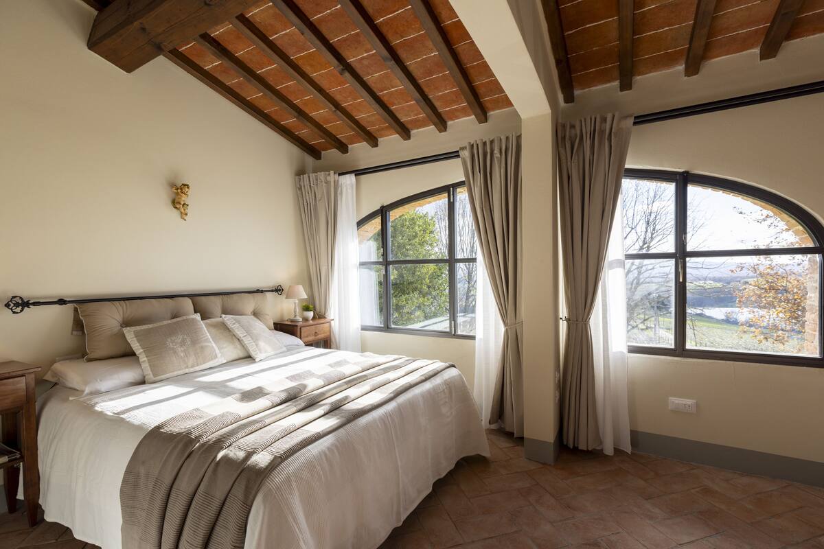 A bedroom with a large bed, wooden ceiling beams, a nightstand with a lamp, and a window with curtains.