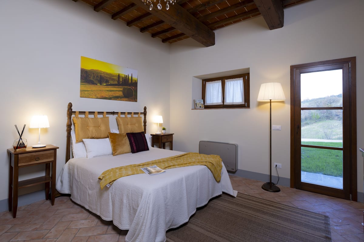 A bedroom with a large bed, wooden headboard, two bedside tables with lamps, a window with curtains, and a view of the outdoors.