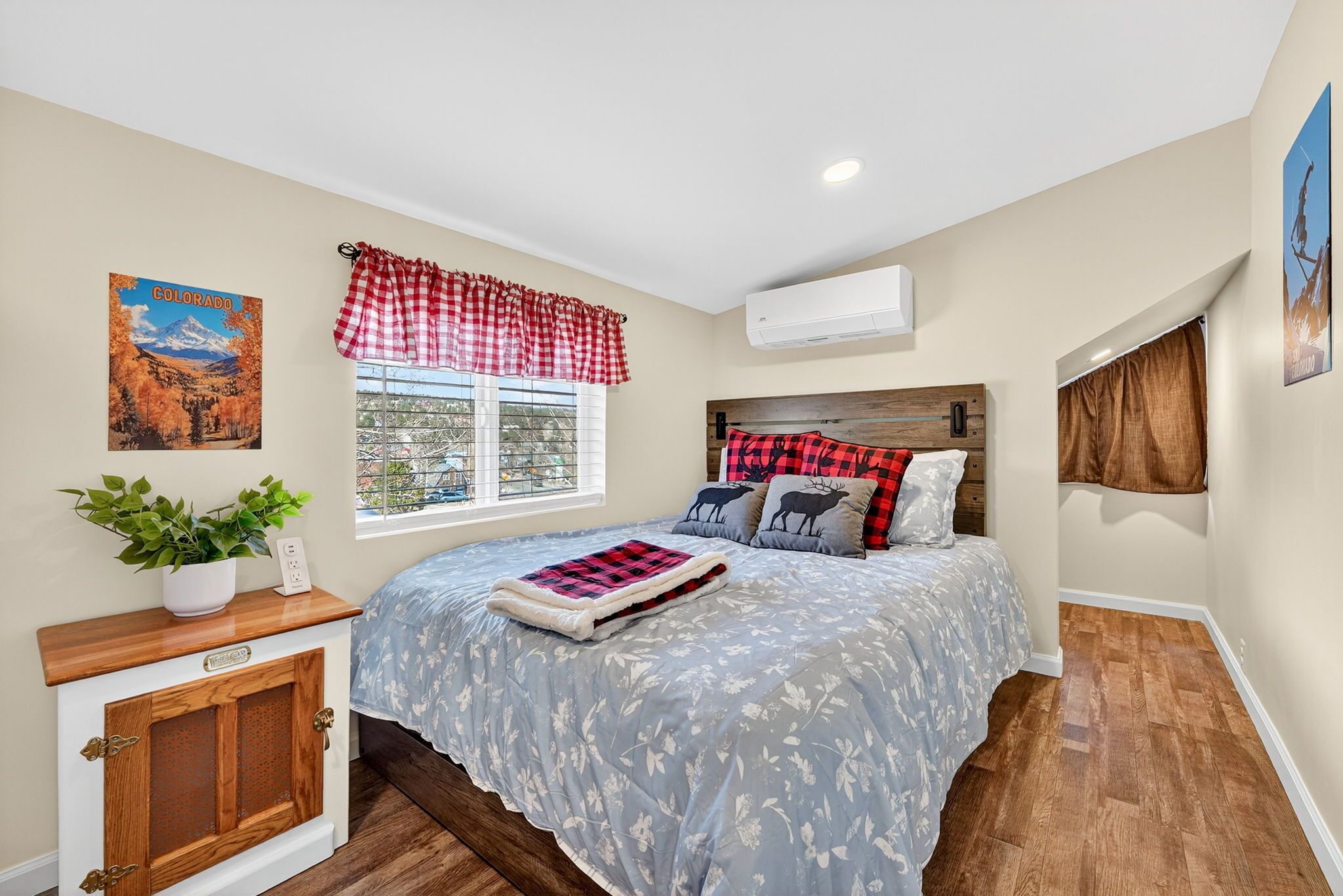 A bedroom with a bed, a window with curtains, a wall-mounted air conditioner, a poster, a small cabinet, and a potted plant.
