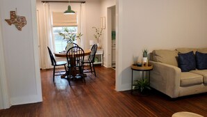 A living room with a beige sofa, a wooden dining table with chairs, a small side table with a plant, and a window with curtains. - Charming Pine Cottage near SFA, azalea gardens,  & walking trails. (Nacogdoches)