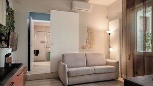 Un salon moderne avec un canapé beige, une table basse et une lampe. On y trouve une porte donnant sur une salle de bain, ainsi qu’un climatiseur fixé au mur.