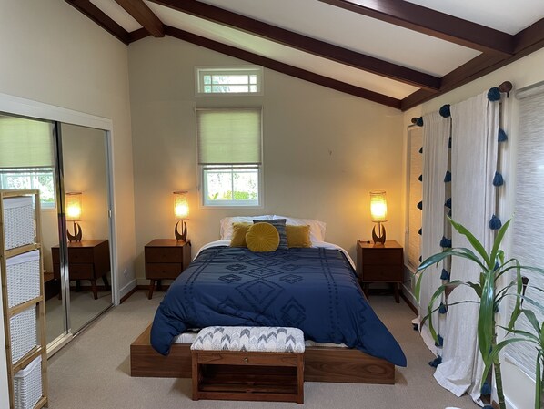 A bedroom with a large bed, two bedside tables with lamps, a window with blinds, a mirror, and a potted plant. - Light filled, tree house, surrounded by nature. 7 minute walk from downtown. (Fairfax)