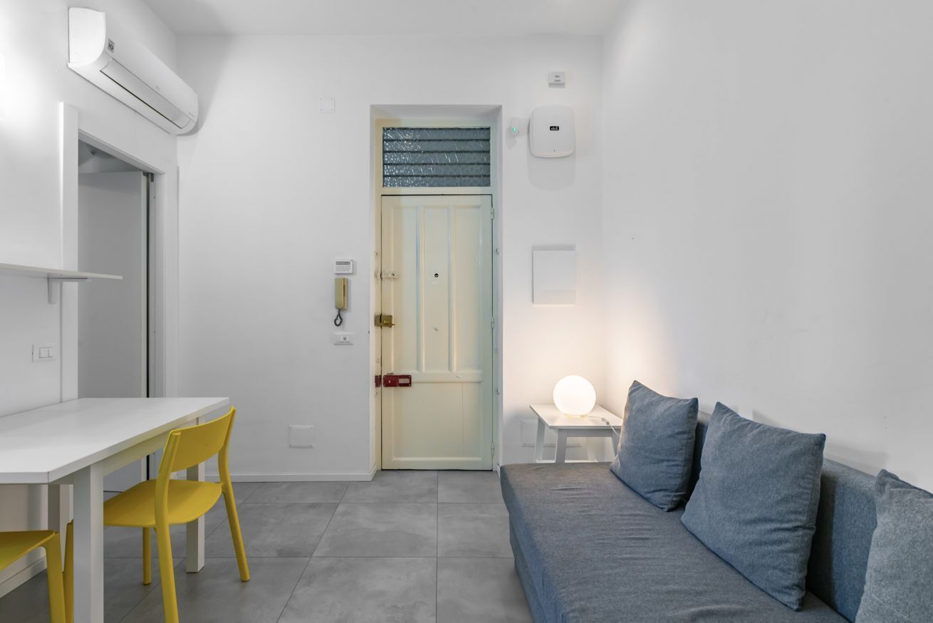 Une pièce de taille modeste, avec une table blanche, des chaises jaunes, un canapé gris et une porte dotée d’un panneau en verre dépoli.
