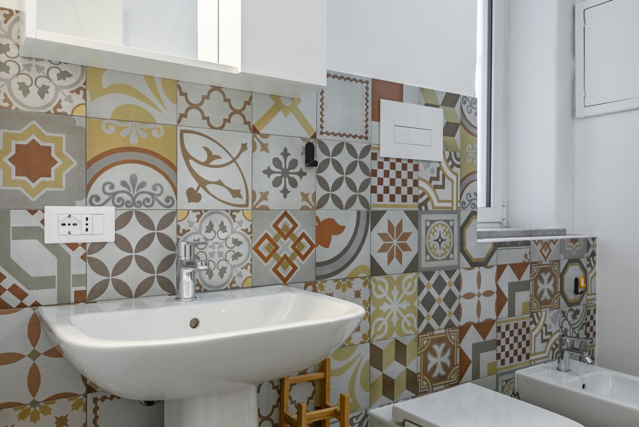 Un baño con un lavabo blanco, un taburete de madera y baldosas estampadas.