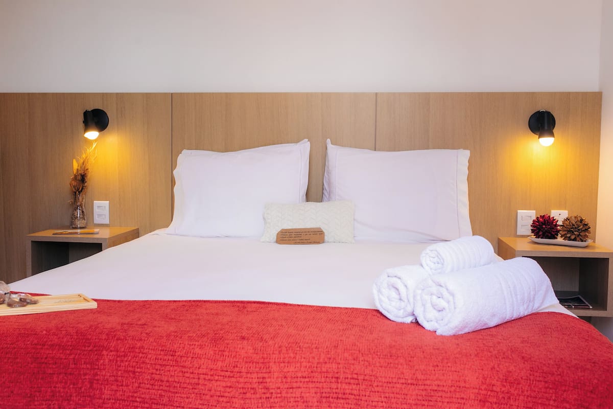 A neatly made bed with white linens, a red blanket, and rolled towels on a wooden headboard.