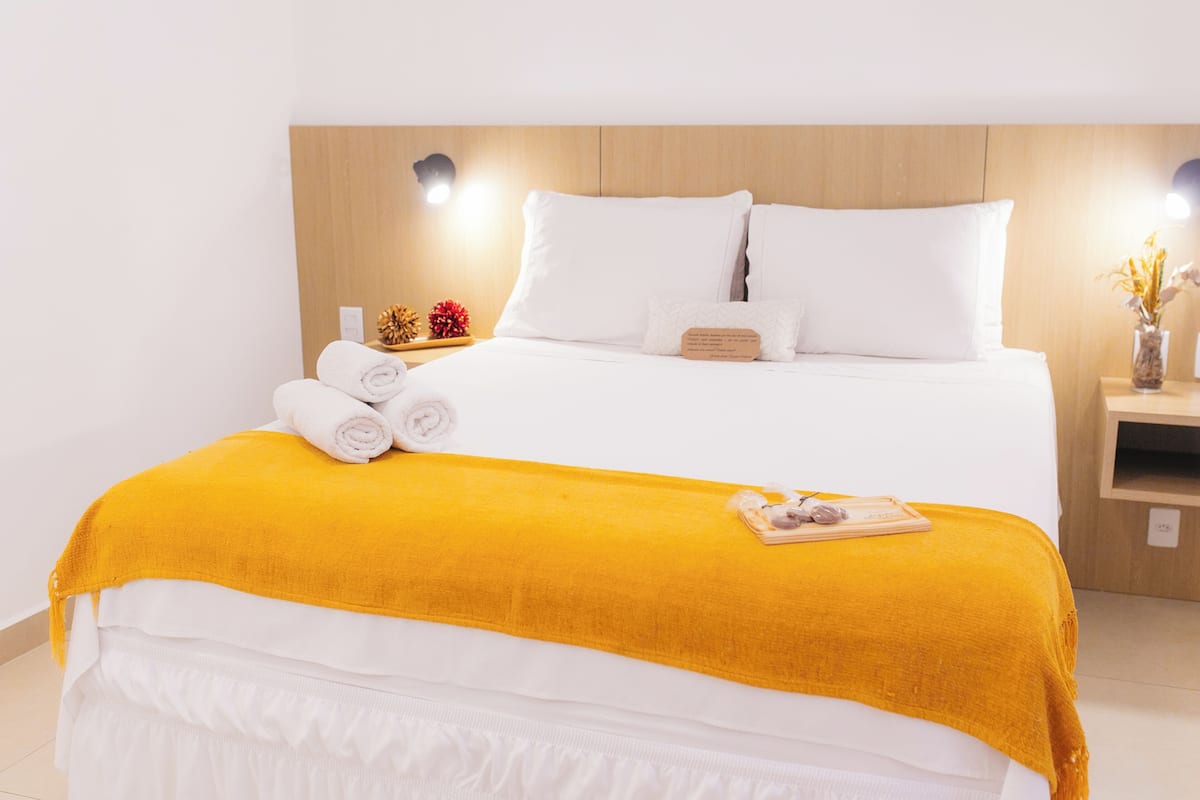 A neatly made bed with white linens and a yellow throw blanket, a bedside table with a book and a small vase, and a wall-mounted light fixture.