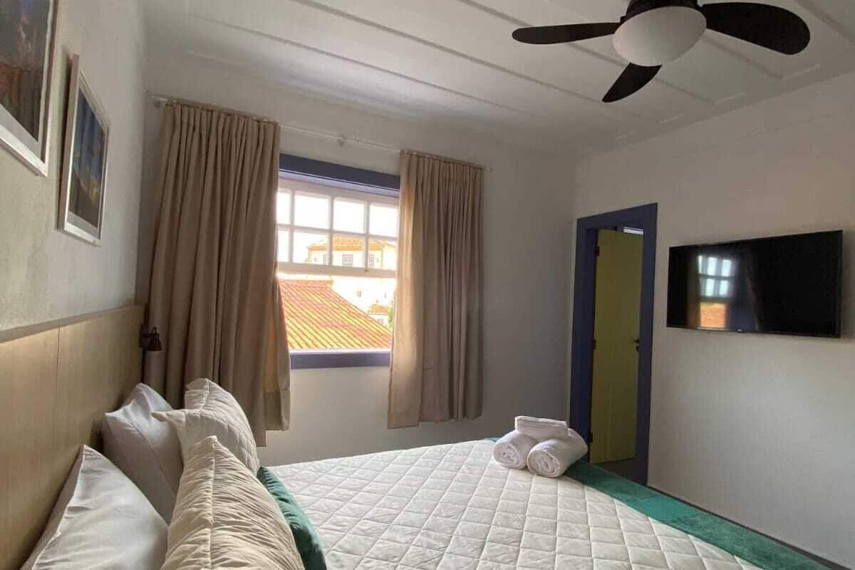 A hotel room with a bed, a ceiling fan, a window with curtains, a flat-screen TV mounted on the wall, and a door.
