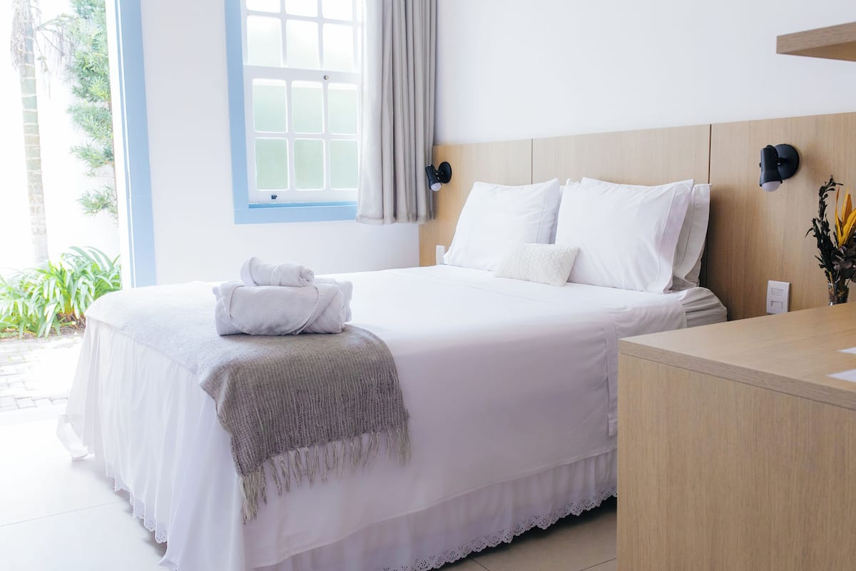 A neatly made bed with white linens and a gray throw blanket, a bedside table with a lamp, and a window with curtains.