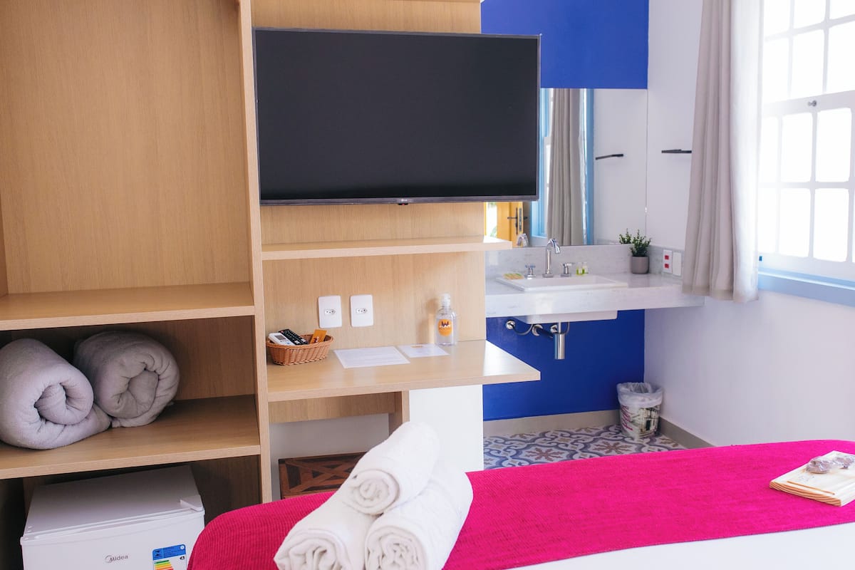 A hotel room with a bed, a TV mounted on a wooden shelf, a sink, and a window with curtains.