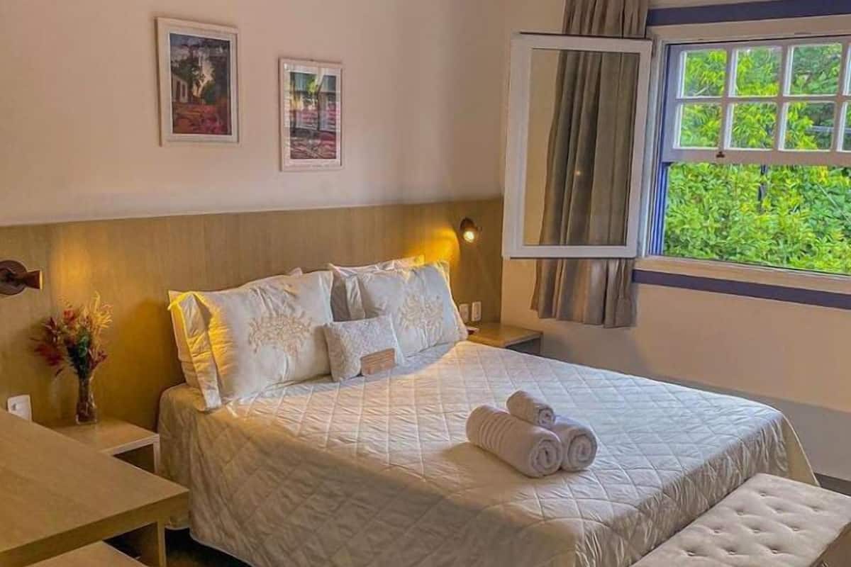 A neatly made bed with a quilted cover, a bedside table with a vase of flowers, and a window with curtains.