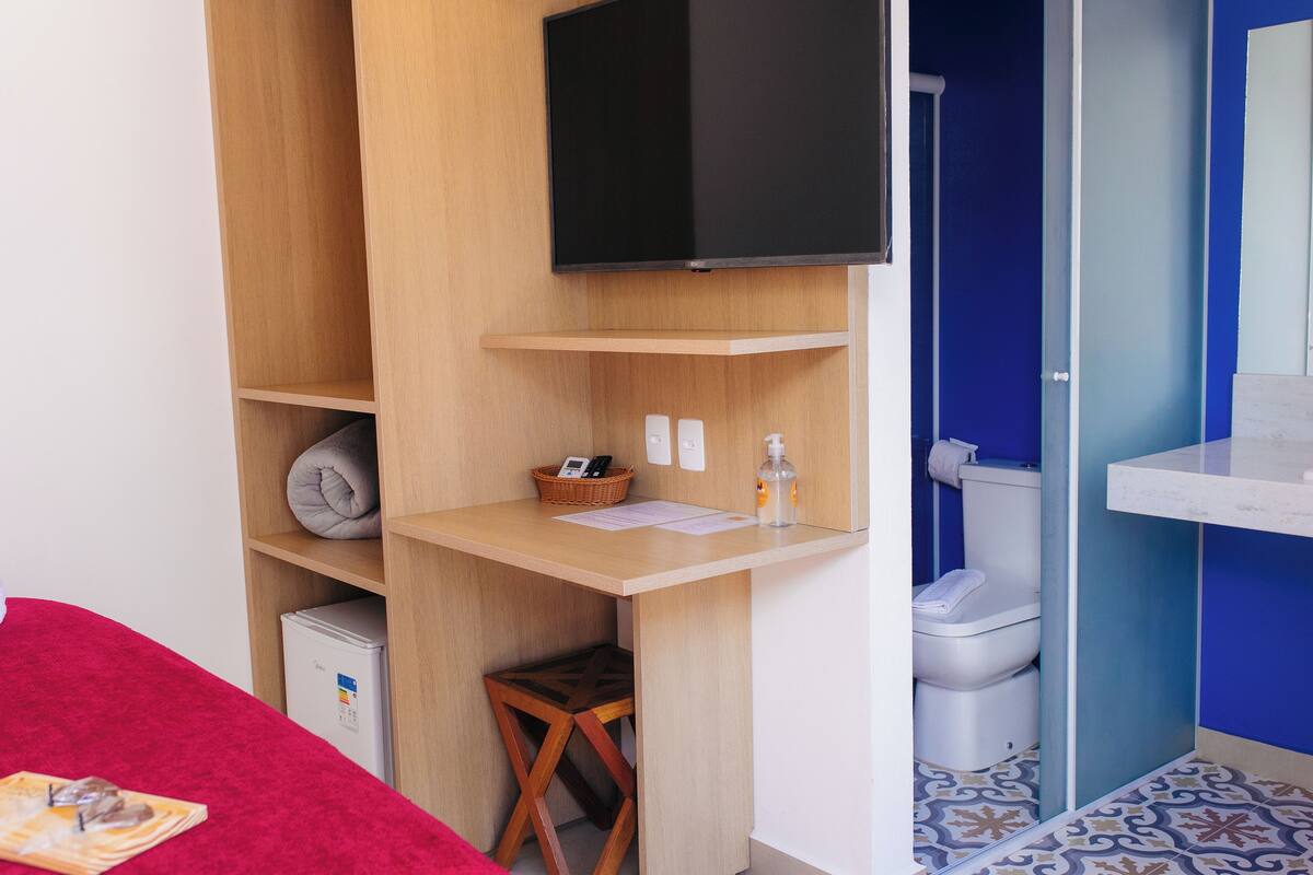 A compact room with a wooden desk, a television, a small refrigerator, and a bathroom with a blue wall and patterned floor.