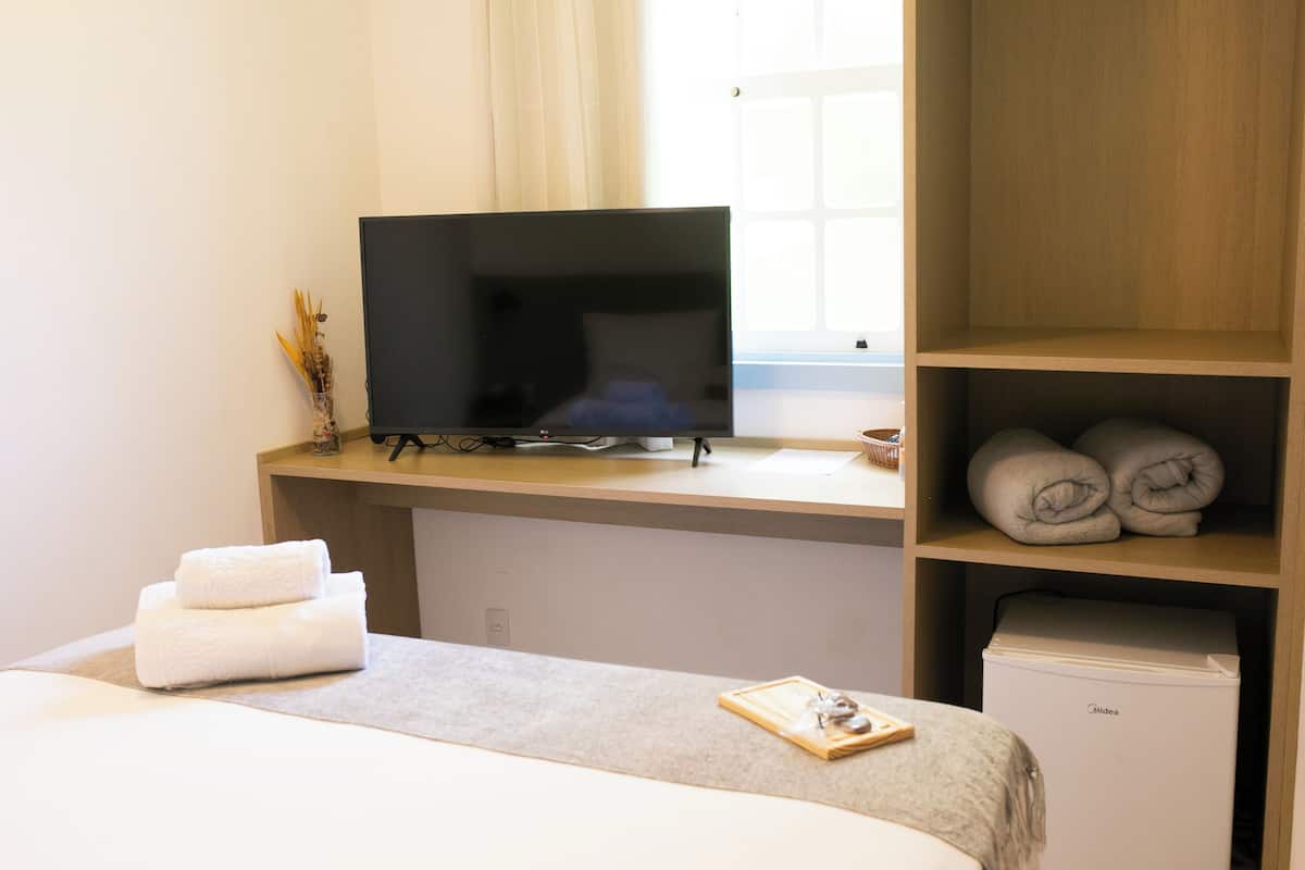 A hotel room with a bed, a television on a stand, a shelf with towels, and a small refrigerator.