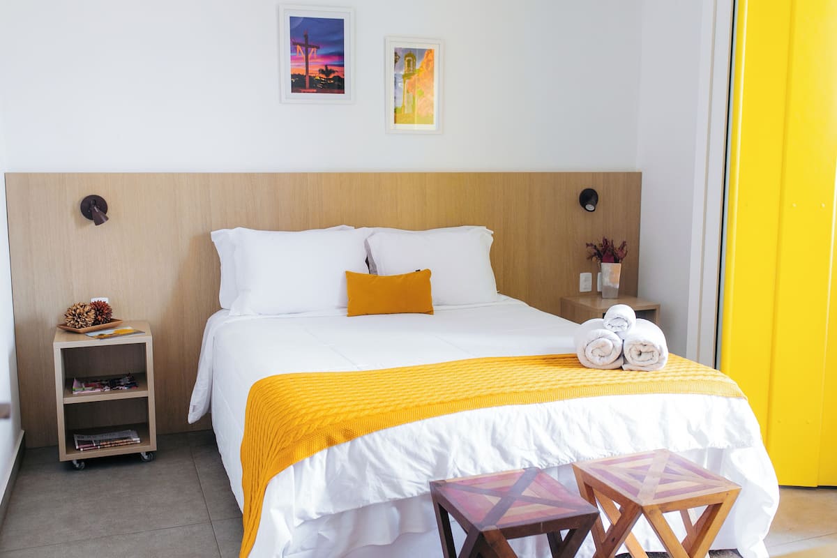 A hotel room with a wooden headboard, a bed with white and yellow bedding, a small wooden nightstand with a lamp, and a small table with towels.