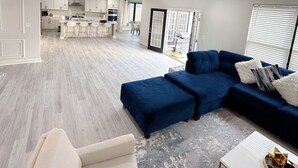 A modern living room with a blue sectional sofa, a white armchair, and a coffee table. There is a kitchen in the background with white cabinets and a dining area. - Embrace Tranquility: A modern retreat home where serenity meets style (Elizabeth City)