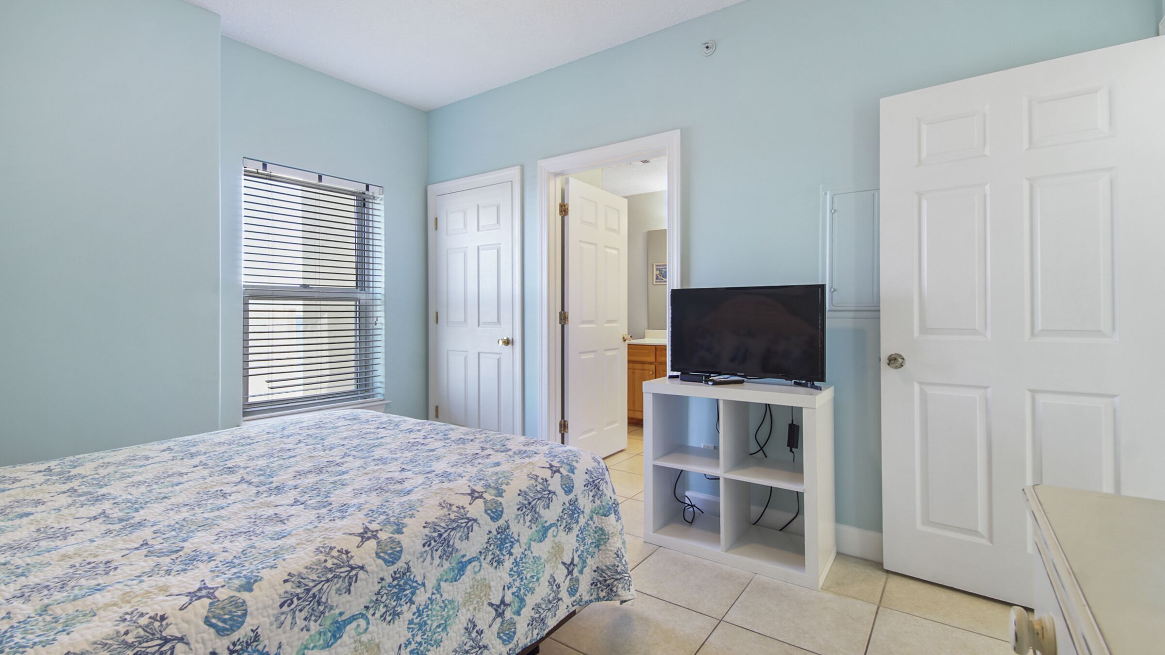 Une chambre à coucher avec un lit, un téléviseur posé sur un meuble, une porte et une fenêtre avec des stores.