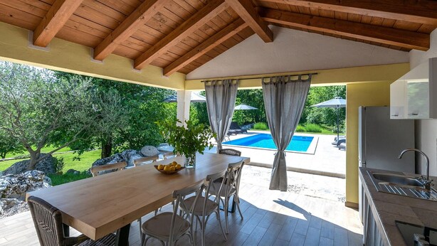Une terrasse couverte avec un plafond en bois, une table à manger avec des chaises, une vue sur une piscine et un coin cuisine avec un évier.