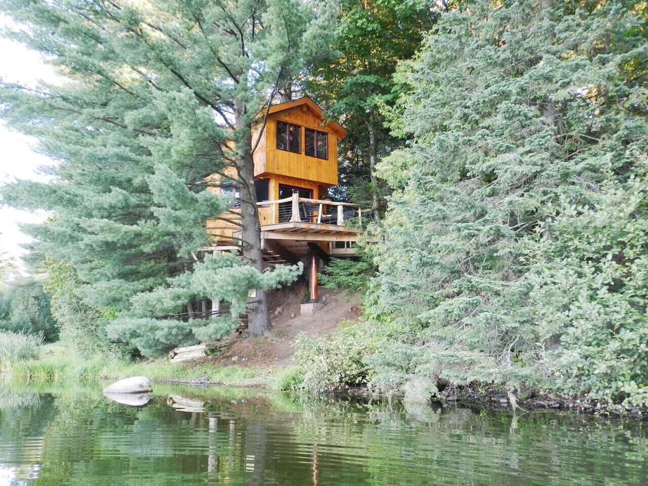 A wooden cabin with a balcony, surrounded by dense trees and a body of water.