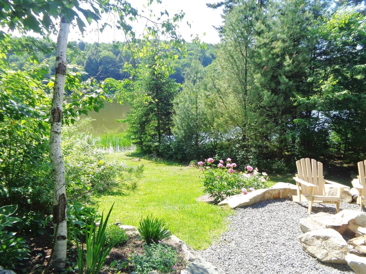 Un jardín con un estanque, rodeado de árboles y arbustos, con sillas Adirondack cerca del agua.