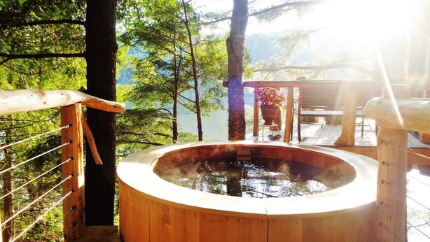 A wooden hot tub surrounded by a wooden deck with a metal railing, set in a forested area with a view of a lake and mountains in the background. - Vermont Tree Cabin on Walker Pond, private retreat (Newport)